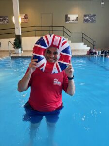 Photo of Logi, the swimming instructor for British Swim School's adaptive swimming lessons. He is standing in an indoor pool holding a circular floatie with the colors of the British flag. He is wearing a red short sleeve shirt and blue shorts.